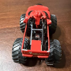 May include: A red monster truck toy with large black tires. The truck has a detailed design, including a front grille and the word "Tasmanian" on the front. The toy is designed for play and collecting.