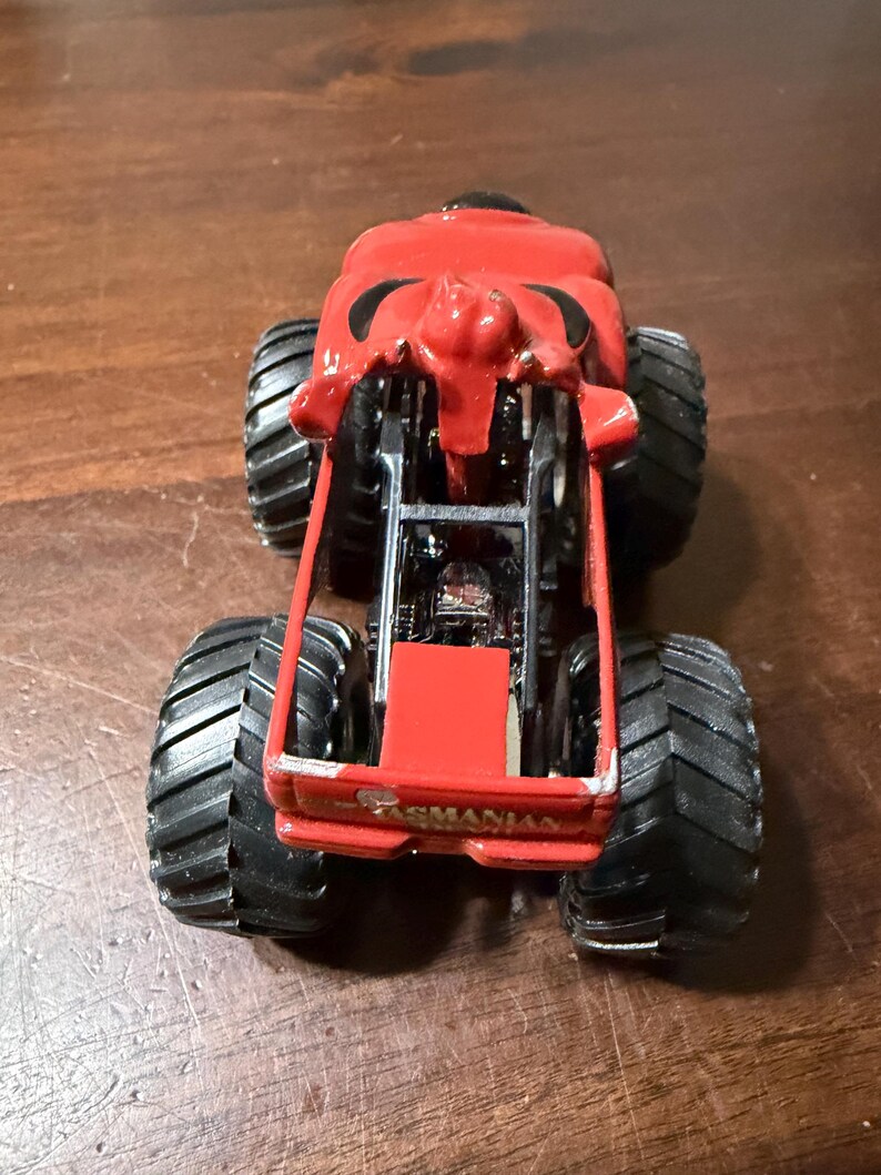 May include: A red monster truck toy with large black tires. The truck has a detailed design, including a front grille and the word "Tasmanian" on the front. The toy is designed for play and collecting.