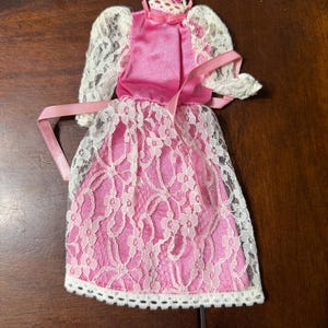 May include: A miniature pink dress with white lace overlay. The dress features a high neckline, short sleeves, and a ribbon sash. A matching lace crown is attached to the top. The dress is designed for a doll.