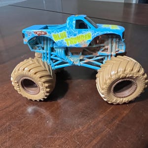 May include: A blue and yellow toy monster truck with large tan tyres. The truck has the words "BLUE THUNDER" on the side. The truck is sitting on a brown surface.