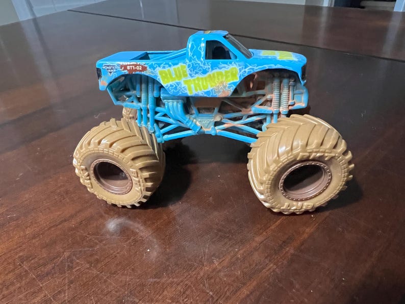 May include: A blue and yellow toy monster truck with large tan tyres. The truck has the words "BLUE THUNDER" on the side. The truck is sitting on a brown surface.