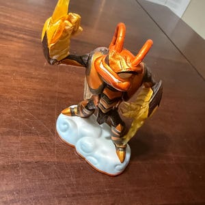 May include: A detailed figurine of a character with an insect-like appearance, featuring orange, brown, and gold accents. The figure is posed dynamically, standing on a cloud-shaped base. The character has large horns and wings.