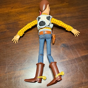 May include: A Woody action figure, viewed from the back, with a brown head and a red bandana. He wears a yellow plaid shirt, a cow-print vest, blue jeans, a brown belt, and brown cowboy boots with yellow spurs. A toy gun holster is attached to the belt.