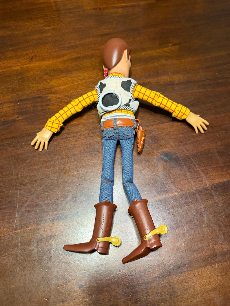 May include: A Woody action figure, viewed from the back, with a brown head and a red bandana. He wears a yellow plaid shirt, a cow-print vest, blue jeans, a brown belt, and brown cowboy boots with yellow spurs. A toy gun holster is attached to the belt.