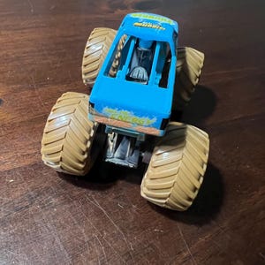May include: A blue monster truck toy with large tan tyres. The truck has the word "ENERGY" printed on the side. The truck is sitting on a wooden surface.