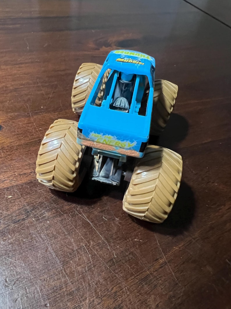 May include: A blue monster truck toy with large tan tyres. The truck has the word "ENERGY" printed on the side. The truck is sitting on a wooden surface.