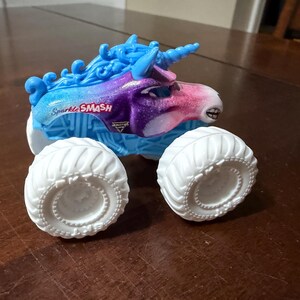 May include: A toy monster truck with a unicorn design. The truck has a blue and purple body with the text "Sparkle SMASH". It has large white tyres and a blue horn.