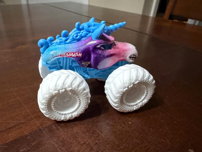 May include: A toy monster truck with a unicorn design. The truck has a blue and purple body with the text "Sparkle SMASH". It has large white tyres and a blue horn.