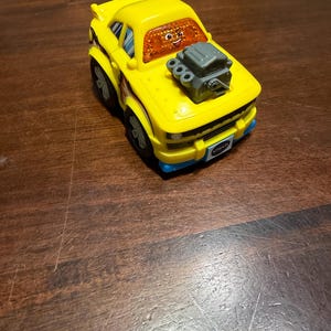 May include: A yellow toy car with black and blue accents. The car has a cartoonish design, featuring a smiling face on the windshield and a gray engine on the hood. The wheels are black, and the car has the brand name "Vtech" on the front.