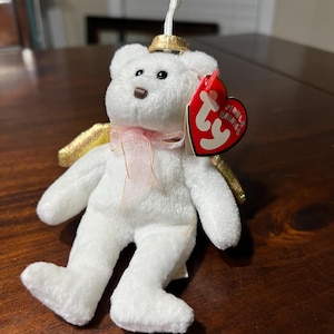 Puede incluir: Un oso de peluche blanco con un lazo de cinta rosa, alas doradas y un halo dorado. El oso tiene ojos negros y una nariz marrón. Una etiqueta roja en forma de corazón está unida al oso.