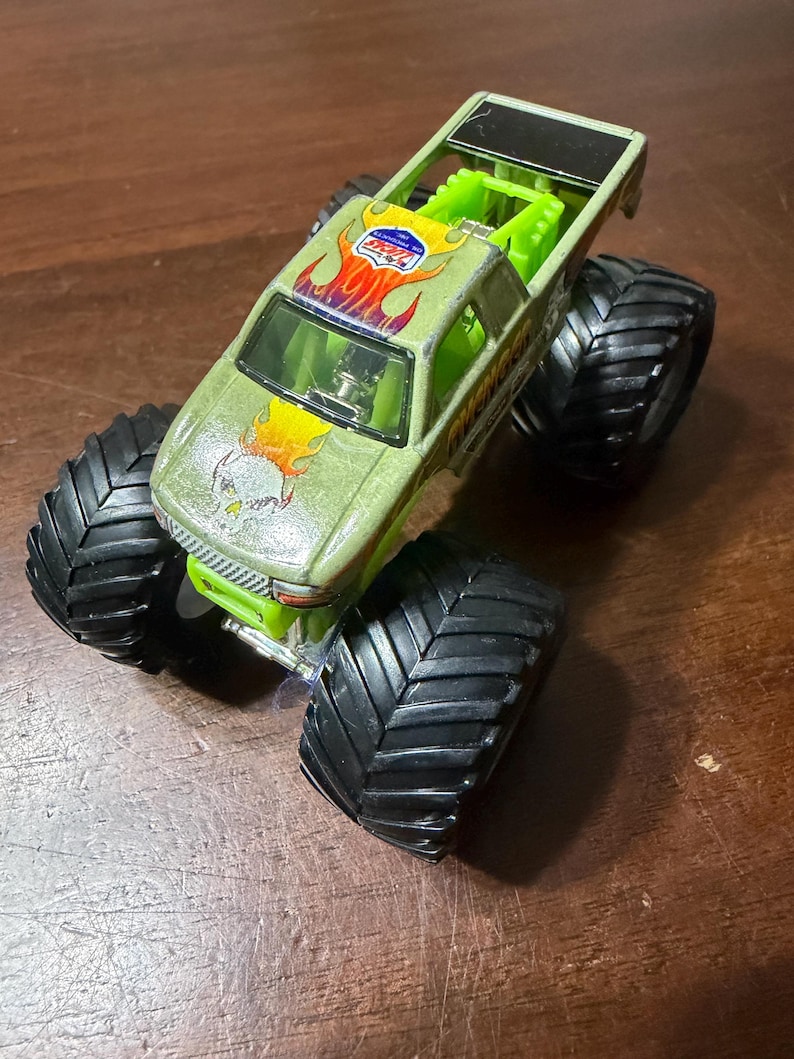 Puede incluir: Un juguete de cami&oacute;n monstruo verde con neum&aacute;ticos negros grandes. El cami&oacute;n presenta un dise&ntilde;o de calavera y llamas en el cap&oacute; y un logotipo en el techo. El cami&oacute;n tiene una jaula antivuelco verde y una parte superior negra.