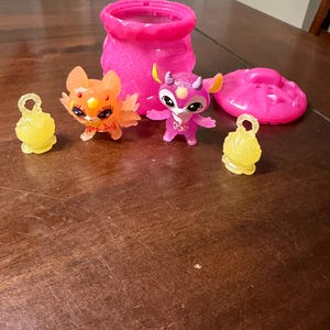 Puede incluir: Colección de juguetes coloridos y brillantes, que incluyen dos pequeñas figuras de criaturas con alas y cuernos, un recipiente rosa, una tapa rosa en forma de platillo y dos amuletos amarillos en forma de campana. Los juguetes están sobre una superficie de madera.
