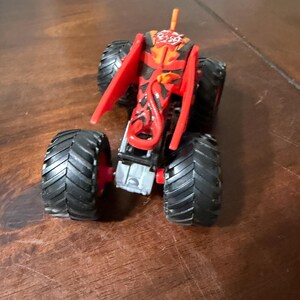 May include: A toy monster truck with large black tires and red accents. The truck's body is primarily red with black and orange flame-like designs. It has a small orange antenna.
