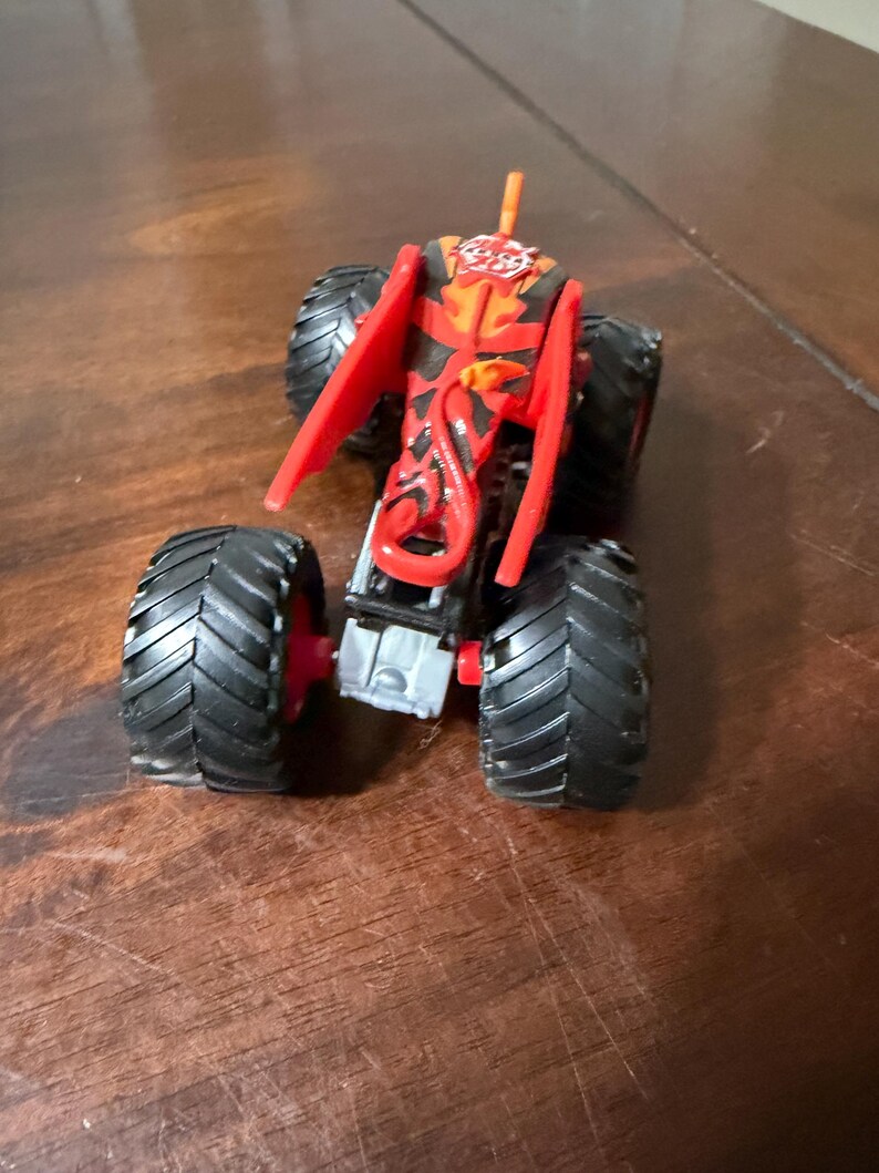 May include: A toy monster truck with large black tires and red accents. The truck's body is primarily red with black and orange flame-like designs. It has a small orange antenna.