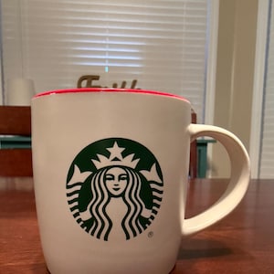 Puede incluir: Taza de cerámica blanca con borde interior rojo y un gran logotipo verde de Starbucks. El logotipo presenta una sirena con cabello suelto y una corona. La taza tiene un asa curva y está sobre una superficie de madera.