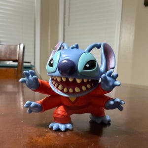 May include: A blue Stitch figurine with large ears, a purple nose, and a wide grin revealing many teeth. The figure is wearing an orange jumpsuit and has four arms and legs. The figurine is posed with its arms outstretched.
