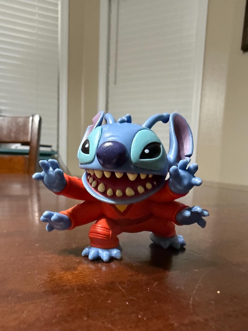 May include: A blue Stitch figurine with large ears, a purple nose, and a wide grin revealing many teeth. The figure is wearing an orange jumpsuit and has four arms and legs. The figurine is posed with its arms outstretched.