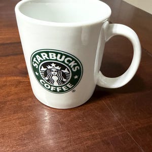 May include: White ceramic coffee mug with a curved handle. The mug features the Starbucks logo in green and black on the front. The mug is sitting on a brown wooden surface.