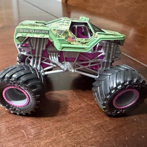 May include: A toy monster truck with large black tires and pink accents. The truck's body is green with the words "SOLDIER FORTUNE" printed on the side. The truck has a camouflage design and a small American flag.