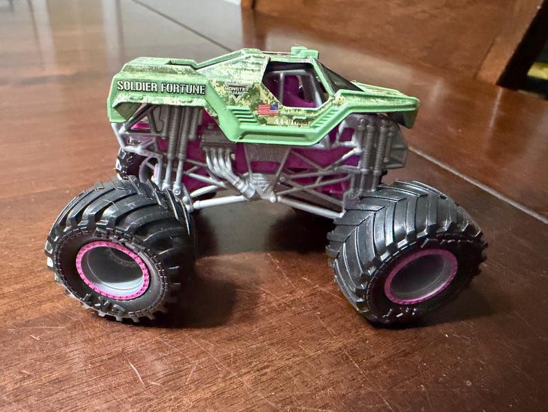 May include: A toy monster truck with large black tires and pink accents. The truck's body is green with the words "SOLDIER FORTUNE" printed on the side. The truck has a camouflage design and a small American flag.