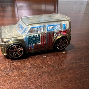 May include: A toy car with a boxy design, in a metallic gold finish. The car has a patriotic theme with a blue and red American flag design on the side. The wheels are black with silver accents.