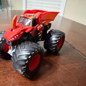 May include: A red and black monster truck toy with large black tires and red rims. The truck has a dragon-like design on the front and top, with orange accents. The toy is sitting on a brown surface.