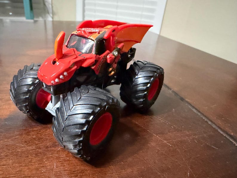 May include: A red and black monster truck toy with large black tires and red rims. The truck has a dragon-like design on the front and top, with orange accents. The toy is sitting on a brown surface.
