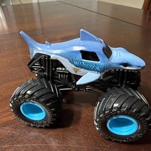 K&ouml;nnte beinhalten: Ein blau-wei&szlig;es Spielzeug-Monstertruck in Form eines Hais, mit dem Wort "MEGALODON" auf der Seite. Der Truck hat gro&szlig;e schwarze Reifen mit blauen Felgen und ein schwarzes Fahrgestell.