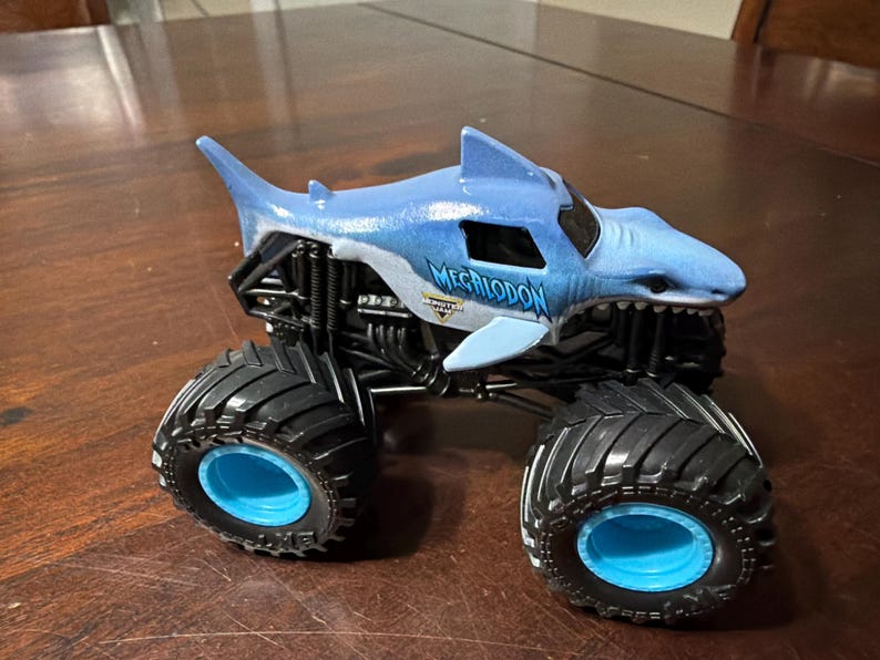K&ouml;nnte beinhalten: Ein blau-wei&szlig;es Spielzeug-Monstertruck in Form eines Hais, mit dem Wort "MEGALODON" auf der Seite. Der Truck hat gro&szlig;e schwarze Reifen mit blauen Felgen und ein schwarzes Fahrgestell.
