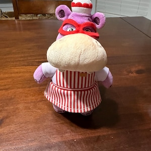 May include: A plush hippo toy wearing a red mask, chef's hat, and apron. The hippo is purple with a tan snout and is wearing a white shirt. The apron has red and white vertical stripes.