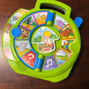 Könnte beinhalten: Ein grünes Fisher-Price-Spielzeug mit Griff, das ein rotierendes Rad mit Tierillustrationen und ein zentrales Bild eines Kindes aufweist. Das Spielzeug enthält Knöpfe und einen Lautsprecher, die für interaktives Spielen und Lernen konzipiert sind.