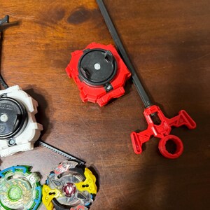 May include: A collection of colorful spinning tops and launchers. The tops feature various designs with red, yellow, green, and blue accents. One red launcher with a black handle is also visible.