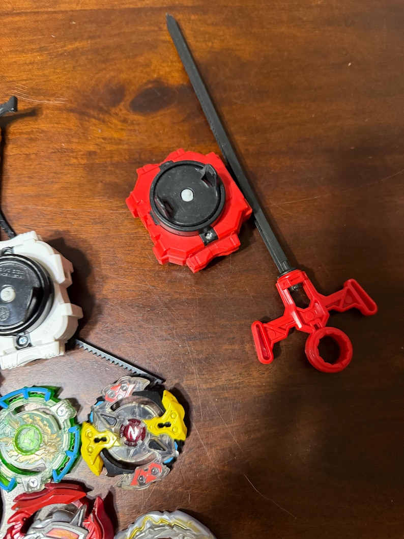 May include: A collection of colorful spinning tops and launchers. The tops feature various designs with red, yellow, green, and blue accents. One red launcher with a black handle is also visible.