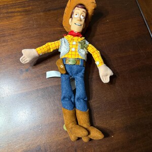 May include: A plush toy of a cowboy character, wearing a brown hat, yellow plaid shirt, blue jeans, and brown boots. The toy has a red neckerchief and a sheriff's badge. The toy's arms are outstretched.