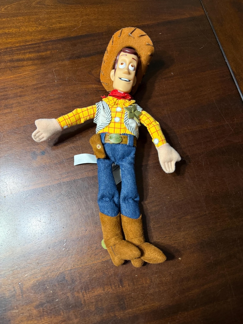 May include: A plush toy of a cowboy character, wearing a brown hat, yellow plaid shirt, blue jeans, and brown boots. The toy has a red neckerchief and a sheriff's badge. The toy's arms are outstretched.