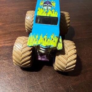 May include: A blue and green toy monster truck with large brown tires. The truck has a graphic of a skull and flames, with the word "Digger" and the year "1996" on the top.