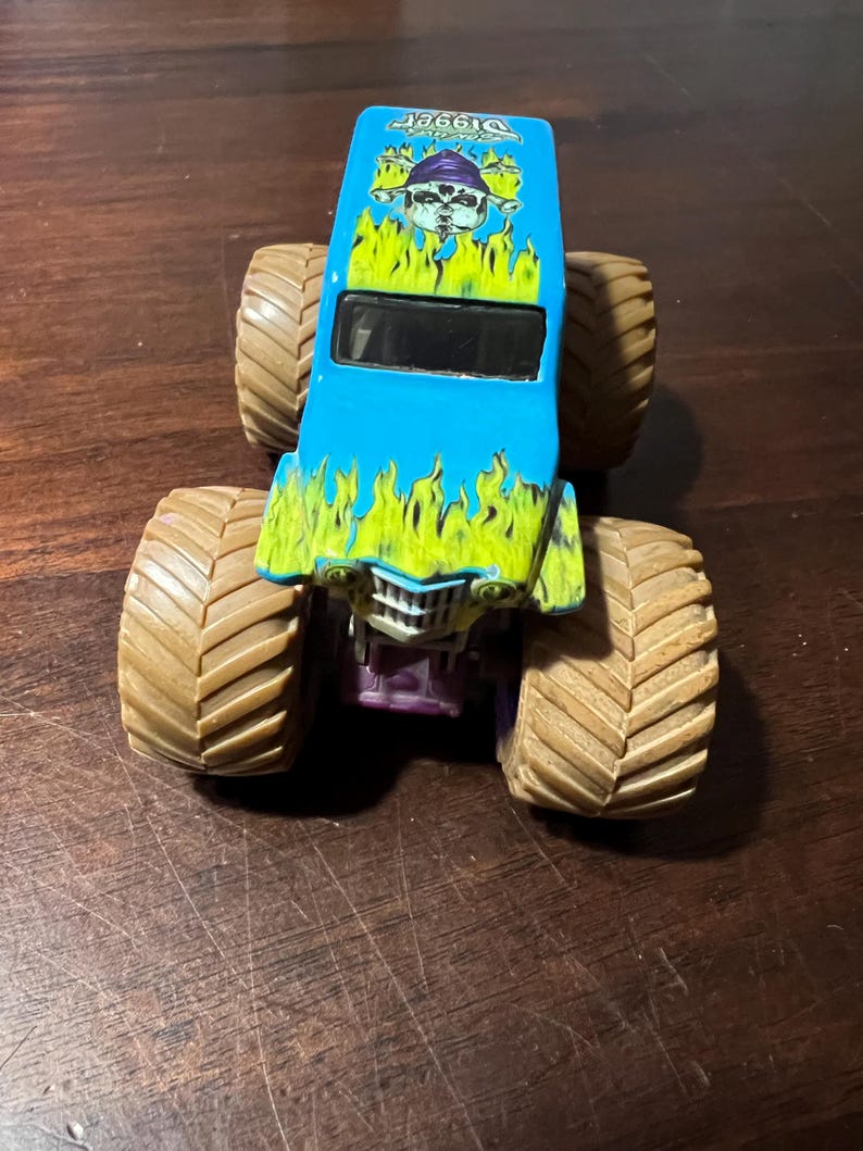 May include: A blue and green toy monster truck with large brown tires. The truck has a graphic of a skull and flames, with the word "Digger" and the year "1996" on the top.