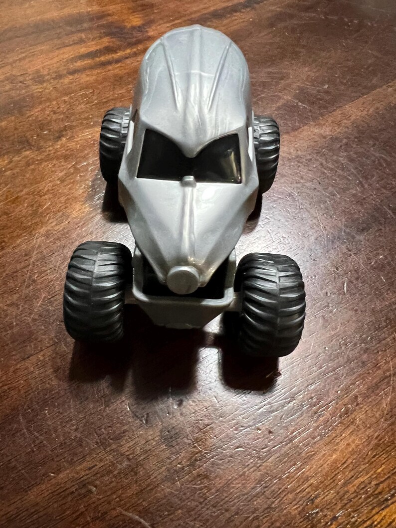 May include: A silver toy car with black tyres. The car has a futuristic design with a rounded body and a large windscreen.