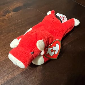 Puede incluir: Un juguete de peluche de vaca roja con manchas blancas y pezuñas blancas. El juguete tiene una etiqueta en forma de corazón Ty adjunta. La vaca está sobre una superficie marrón oscuro.