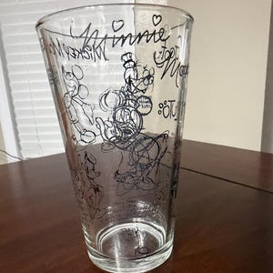 May include: A clear glass tumbler featuring black line art illustrations of various cartoon characters, including Mickey Mouse and Goofy. The design includes the text "Minnie" and "Mickey Mouse" written in a script font, along with small heart icons.
