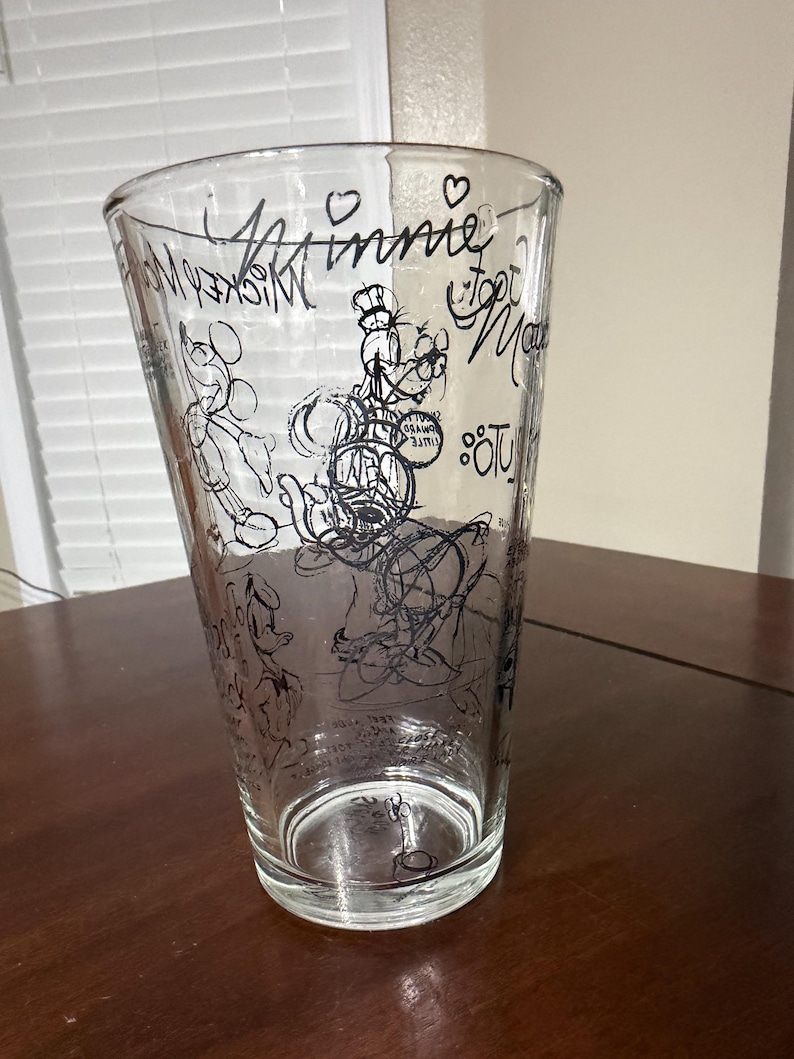 May include: A clear glass tumbler featuring black line art illustrations of various cartoon characters, including Mickey Mouse and Goofy. The design includes the text "Minnie" and "Mickey Mouse" written in a script font, along with small heart icons.