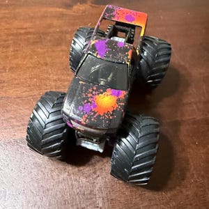 Pode incluir: Um brinquedo de caminh&atilde;o monstro preto com pneus pretos grandes. A carroceria do caminh&atilde;o &eacute; decorada com respingos de tinta roxa, laranja e amarela. A parte superior do caminh&atilde;o &eacute; laranja e as rodas s&atilde;o pretas.