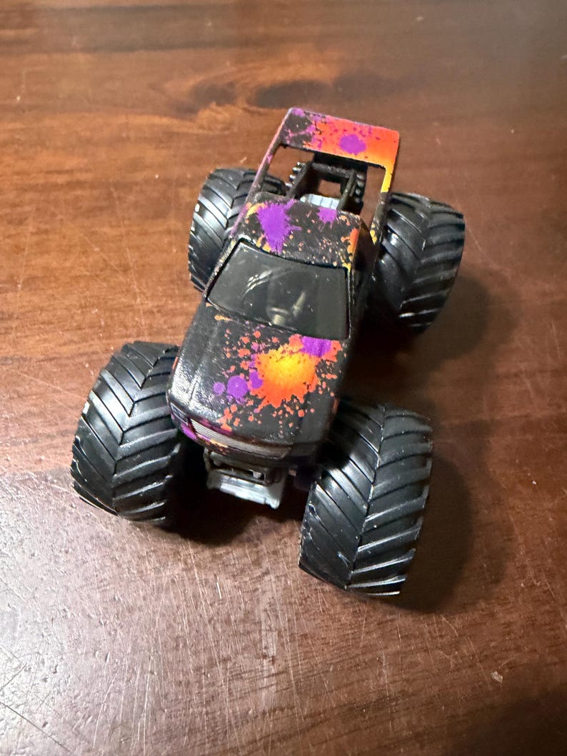 Pode incluir: Um brinquedo de caminh&atilde;o monstro preto com pneus pretos grandes. A carroceria do caminh&atilde;o &eacute; decorada com respingos de tinta roxa, laranja e amarela. A parte superior do caminh&atilde;o &eacute; laranja e as rodas s&atilde;o pretas.