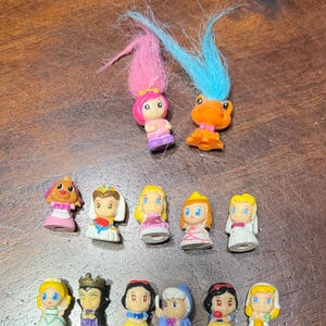 May include: A selection of small, colourful figurines, including troll dolls with pink and blue hair, and various princess figures. The figurines are made of plastic and have detailed features, with a variety of dresses and accessories.