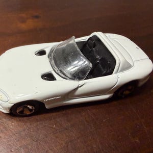Puede incluir: Un coche descapotable de juguete blanco con interior negro y parabrisas transparente. El coche tiene ruedas negras y está sobre una superficie marrón. Es una réplica en miniatura de un coche deportivo.