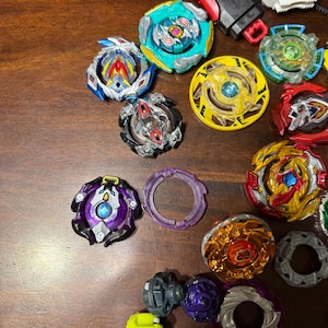 May include: A collection of colorful spinning tops in various designs and colors, including blue, yellow, red, and purple. Some tops have intricate layered designs, while others have simple, solid colors. A red and black launcher is also visible.