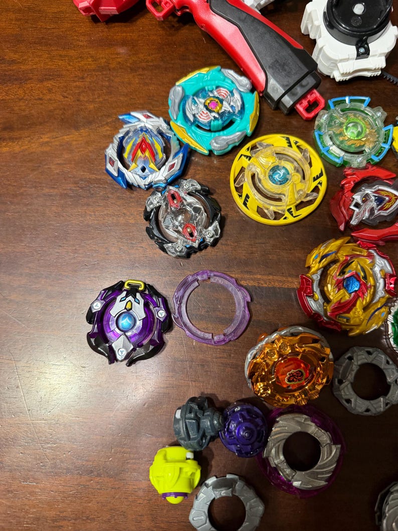 May include: A collection of colorful spinning tops in various designs and colors, including blue, yellow, red, and purple. Some tops have intricate layered designs, while others have simple, solid colors. A red and black launcher is also visible.