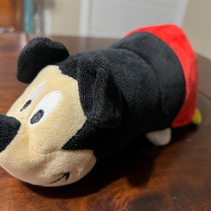 May include: A Mickey Mouse plush toy, with a black body, ears, and nose, a cream-coloured face, and a red tail. The toy is lying on a wooden surface.