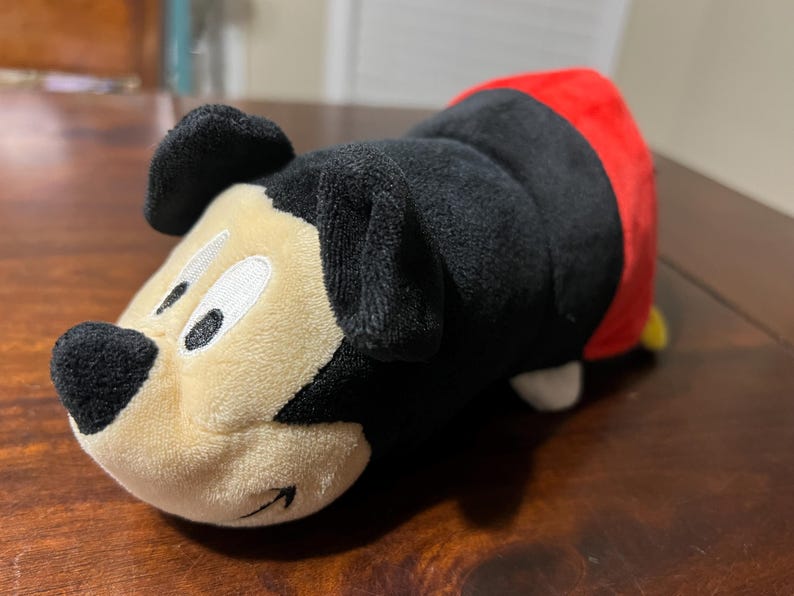 May include: A Mickey Mouse plush toy, with a black body, ears, and nose, a cream-coloured face, and a red tail. The toy is lying on a wooden surface.