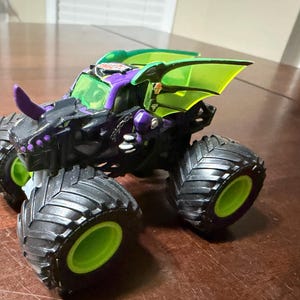 May include: A black, purple, and green monster truck toy with large, black tyres and green rims. The truck features a dragon-themed design with a purple horn and green wing-like accents. The word "Dragonoidz" is visible on the side.